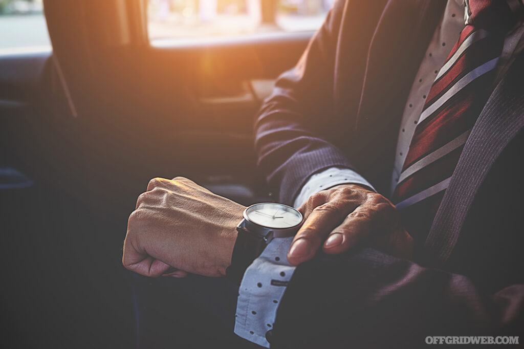 Photo of a formally dressed man anxiously checking his watch in the back seat of a car while trying to be cool about his situation.