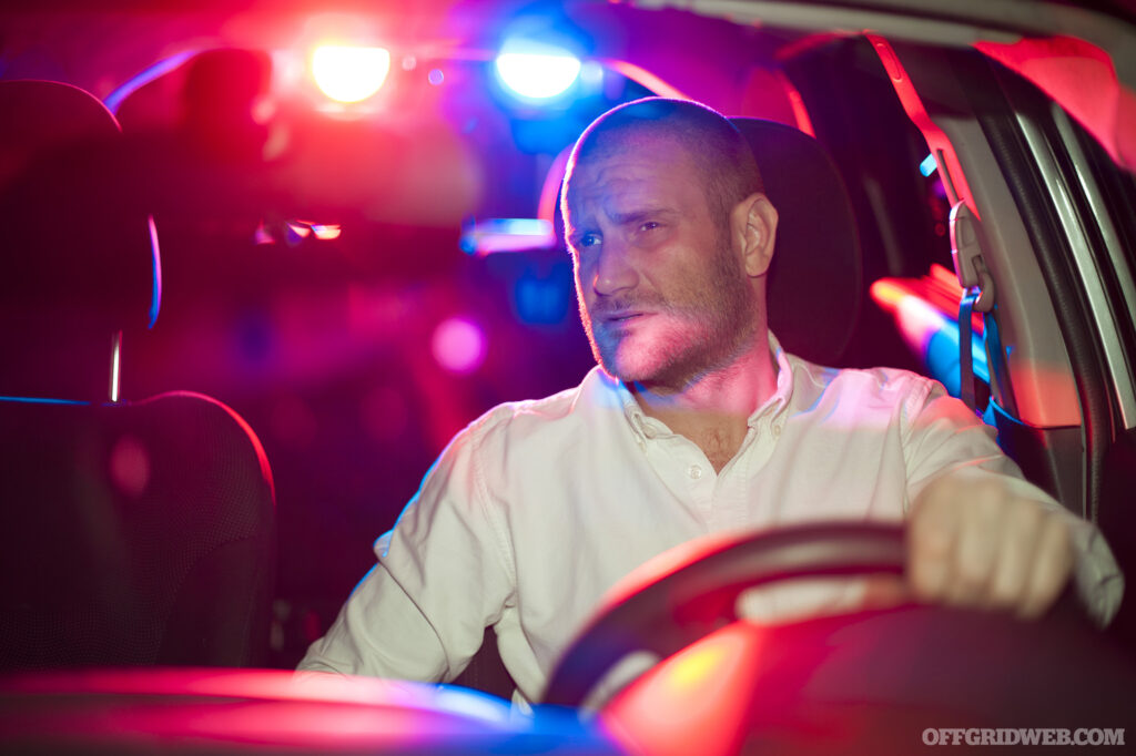 Male driver being pulled over. A police patrol cruiser is visible in the rear window with its red and blue emergency lights on.