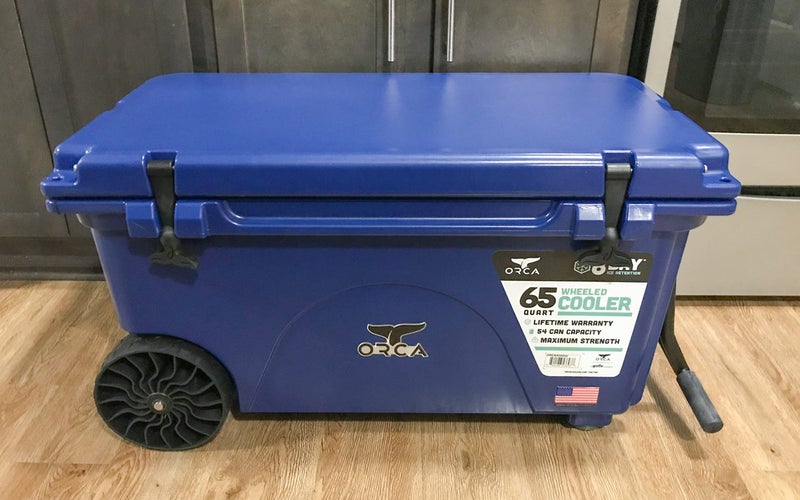 A blue ORCA 65-Quart Insulated Rolling Cooler sits on the floor in front of some kitchen cabinets.