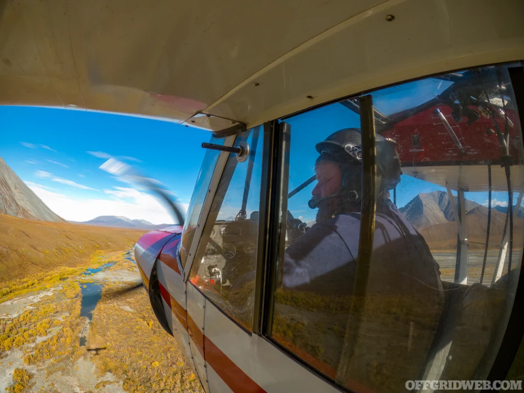 Photo of a man from the outside looking in, flying a bush plane over Alaskan wilderness.