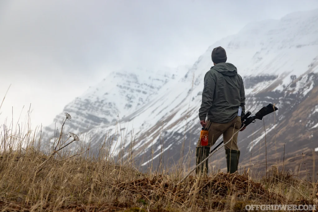 Photo of an adult male standing on a grassy foothill looking at snow covered mountains in the distance.
